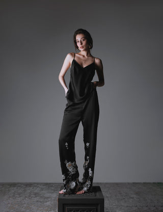 A woman modeling a matching silk set with a black-based print, consisting of a sleeveless top and long pants. She is standing on a display cube in front of a dark, moody background.