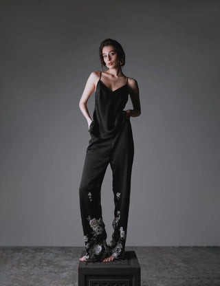 A woman modeling a matching silk set with a black-based print, consisting of a sleeveless top and long pants. She is standing on a display cube in front of a dark, moody background.