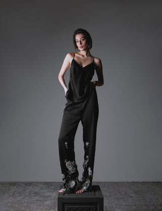 Woman wearing a black silk set composed by long pants and a v neck top with spaghetti straps and with floral patterns on a gray background
