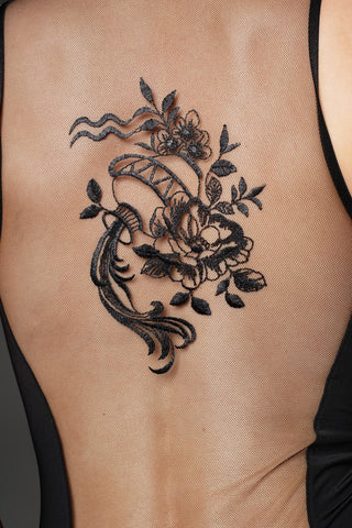 A close-up of a person's bare back with a black floral and scroll tattoo applied over sheer black mesh fabric, with a black strap visible at the shoulder.