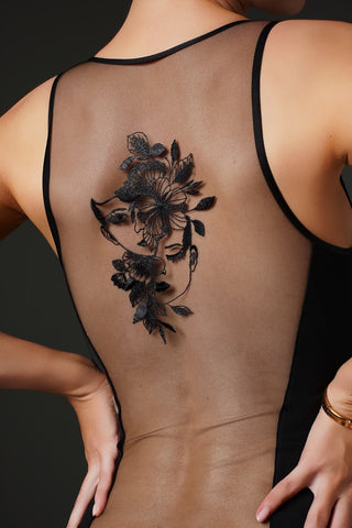 A woman shown from the back wearing a black mesh camisole, featuring a large black floral tattoo on her upper back.