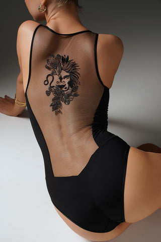 A woman wearing a black one-piece bodysuit with a sheer mesh back panel, photographed from behind. The bodysuit features a large black floral and lion graphic printed on the back, set against a dark gray studio background.