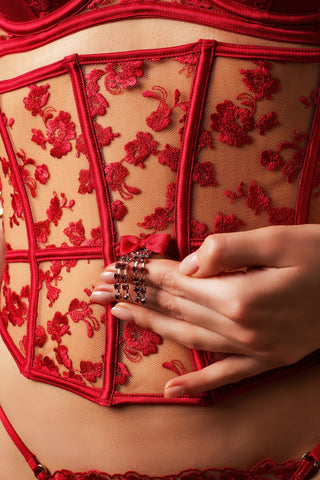Close-up of a person wearing a red lace and mesh corset-style lingerie piece, with a hand resting on the front over a decorative ring.